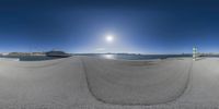 a fisheye lens photo of a beach with snow capped mountains in the distance and people on a bench, with sun reflecting off