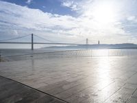 an empty sidewalk sitting on the water with the sun reflecting off the sky and bridge in the distance