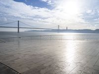 an empty sidewalk sitting on the water with the sun reflecting off the sky and bridge in the distance