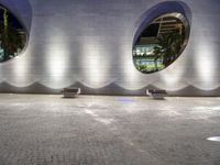 two benches sit on a tiled walkway in front of a building with circular windows above