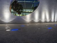 a large building with a circular mirror at night, with the surrounding concretes, has blue lighting from outside