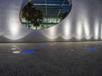 a large building with a circular mirror at night, with the surrounding concretes, has blue lighting from outside