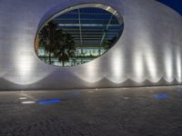 a large building with a circular mirror at night, with the surrounding concretes, has blue lighting from outside