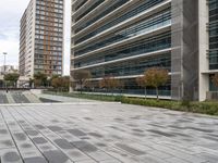 the empty street has grey bricked sidewalks in front of tall buildings and a fenced area