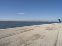 a sandy beach with lots of water and buildings in the background and people standing near the ocean on the shore