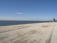 a sandy beach with lots of water and buildings in the background and people standing near the ocean on the shore