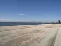 a sandy beach with lots of water and buildings in the background and people standing near the ocean on the shore