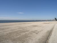 a sandy beach with lots of water and buildings in the background and people standing near the ocean on the shore