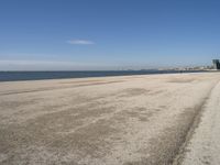 a sandy beach with lots of water and buildings in the background and people standing near the ocean on the shore