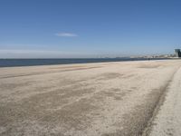 a sandy beach with lots of water and buildings in the background and people standing near the ocean on the shore