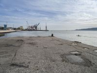 a concrete pier on the edge of the water in a city with cranes in the distance