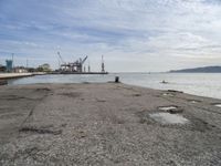 a concrete pier on the edge of the water in a city with cranes in the distance