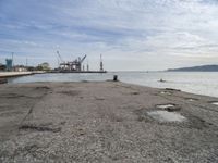 a concrete pier on the edge of the water in a city with cranes in the distance