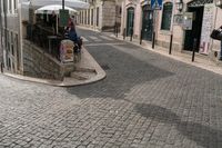 people walking down a cobblestone street in front of a store that has an umbrella over the entrance