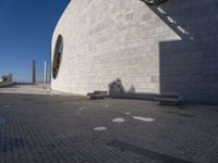 a person sitting on a bench outside a building by the sea and in front of the wall with the shadow on the ground