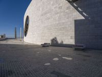 a person sitting on a bench outside a building by the sea and in front of the wall with the shadow on the ground