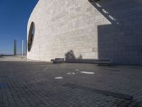 a person sitting on a bench outside a building by the sea and in front of the wall with the shadow on the ground