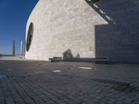 a person sitting on a bench outside a building by the sea and in front of the wall with the shadow on the ground