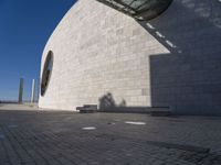 a person sitting on a bench outside a building by the sea and in front of the wall with the shadow on the ground