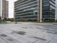 a brick plaza with several trees near buildings and green grass area next to it and empty seating on the sides of the area