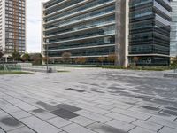 a brick plaza with several trees near buildings and green grass area next to it and empty seating on the sides of the area