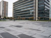 a brick plaza with several trees near buildings and green grass area next to it and empty seating on the sides of the area