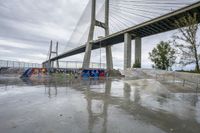 a skate park under a bridge with graffiti on it's wall and in the ground is a river