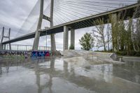 a skate park under a bridge with graffiti on it's wall and in the ground is a river