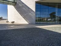 there is an empty bench on the sidewalk in front of the large building with a reflection of it