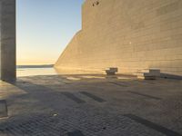 a shadow on an empty concrete area with some benches near by the water and two concrete pillars