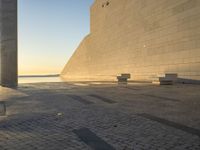 a shadow on an empty concrete area with some benches near by the water and two concrete pillars