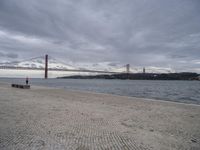 a stone area with benches on the beach in front of a bridge, water and buildings