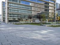 a paved square in front of a large glass building with buildings on both sides and cars driving on the other side