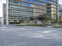 a paved square in front of a large glass building with buildings on both sides and cars driving on the other side