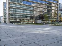 a paved square in front of a large glass building with buildings on both sides and cars driving on the other side