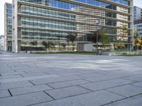 a paved square in front of a large glass building with buildings on both sides and cars driving on the other side