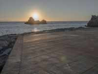 a lone bench on a brick walkway overlooking the ocean at sunset with a rock out in front