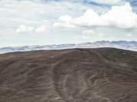 a lone mountain that is standing in the dirt with hills behind it and many clouds