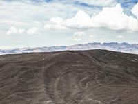 a lone mountain that is standing in the dirt with hills behind it and many clouds