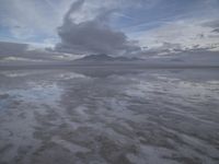 a lonely beach with cloudy skies in the distance, reflecting water on the sand and a building on the shore of the other side