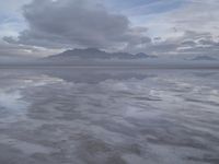 a lonely beach with cloudy skies in the distance, reflecting water on the sand and a building on the shore of the other side