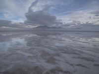 a lonely beach with cloudy skies in the distance, reflecting water on the sand and a building on the shore of the other side