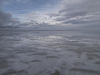 a lonely beach with cloudy skies in the distance, reflecting water on the sand and a building on the shore of the other side