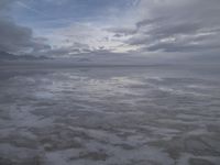 a lonely beach with cloudy skies in the distance, reflecting water on the sand and a building on the shore of the other side