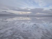 a lonely beach with cloudy skies in the distance, reflecting water on the sand and a building on the shore of the other side