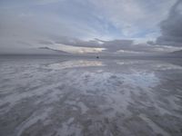a lonely beach with cloudy skies in the distance, reflecting water on the sand and a building on the shore of the other side