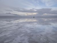 a lonely beach with cloudy skies in the distance, reflecting water on the sand and a building on the shore of the other side