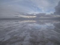 a lonely beach with cloudy skies in the distance, reflecting water on the sand and a building on the shore of the other side