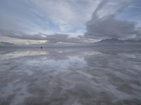 a lonely beach with cloudy skies in the distance, reflecting water on the sand and a building on the shore of the other side