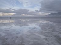 a lonely beach with cloudy skies in the distance, reflecting water on the sand and a building on the shore of the other side